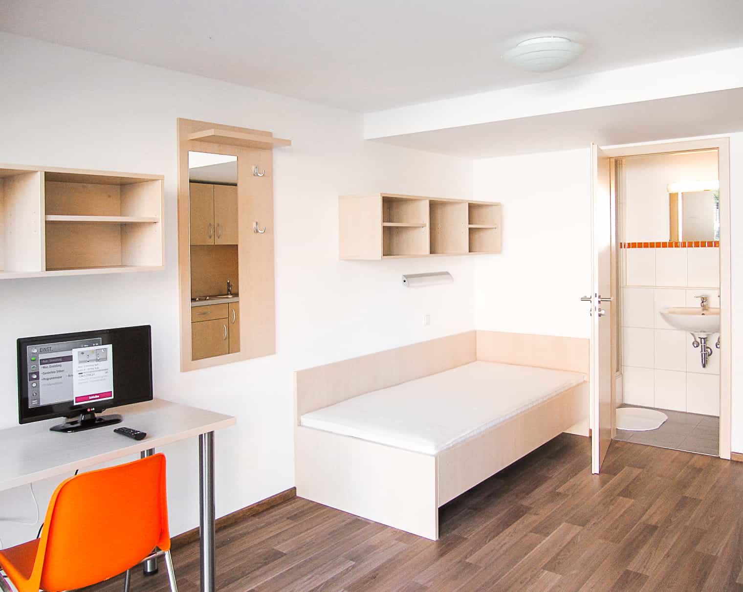 Ein einfaches Einzelzimmer mit einem Bett, Schreibtisch und Waschbecken in einer neutralen Farbgestaltung.