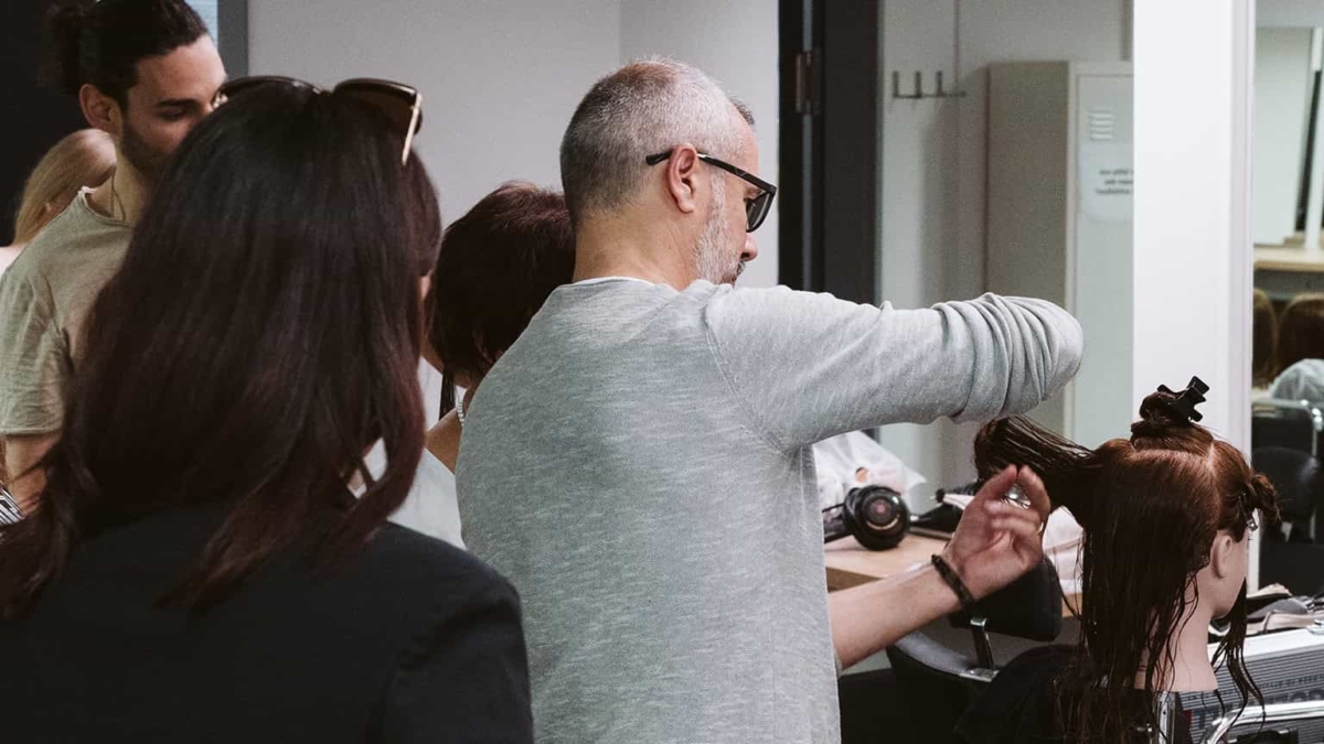 Friseur schneidet Haare eines Kunden in einem modernen Salon mit mehreren Personen im Hintergrund.