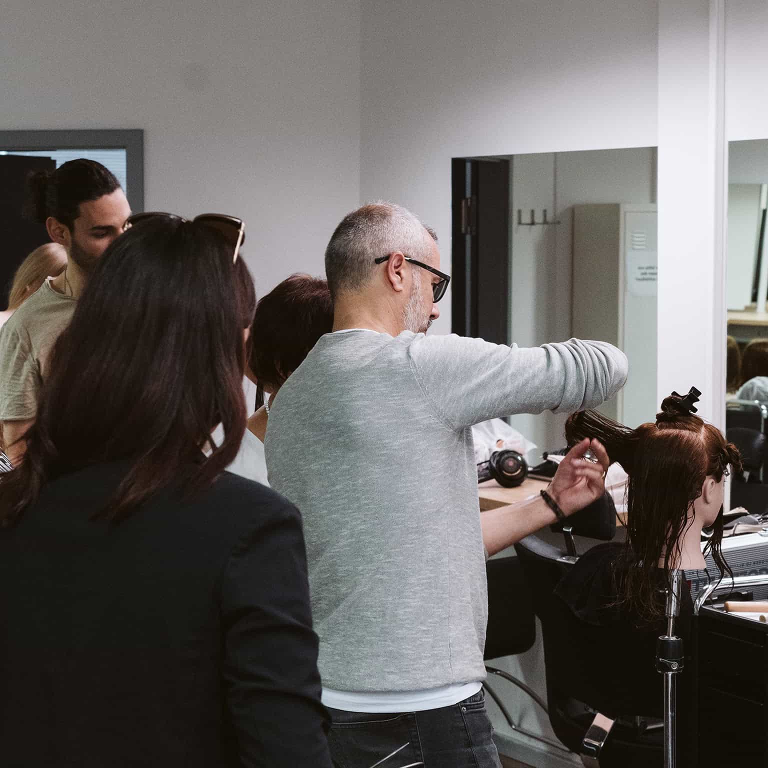 Friseur schneidet Haare eines Kunden in einem modernen Salon mit mehreren Personen im Hintergrund.