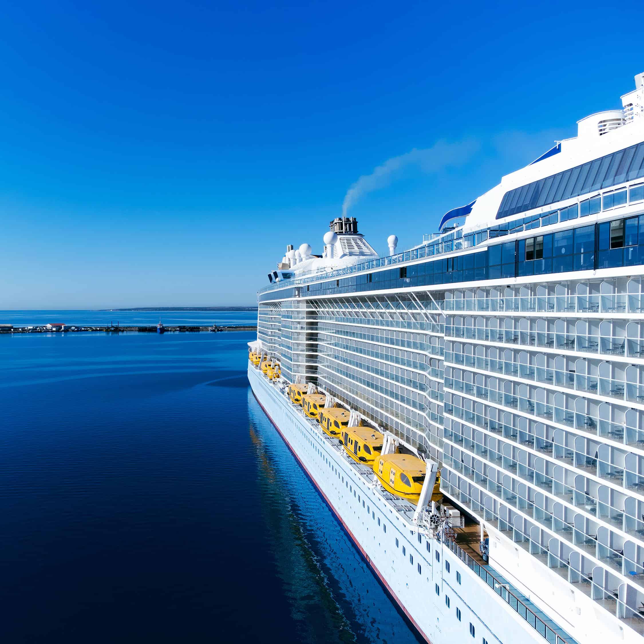 Ein großes Kreuzfahrtschiff mit gelben Rettungsbooten an der Seite im Hafen unter blauem Himmel.