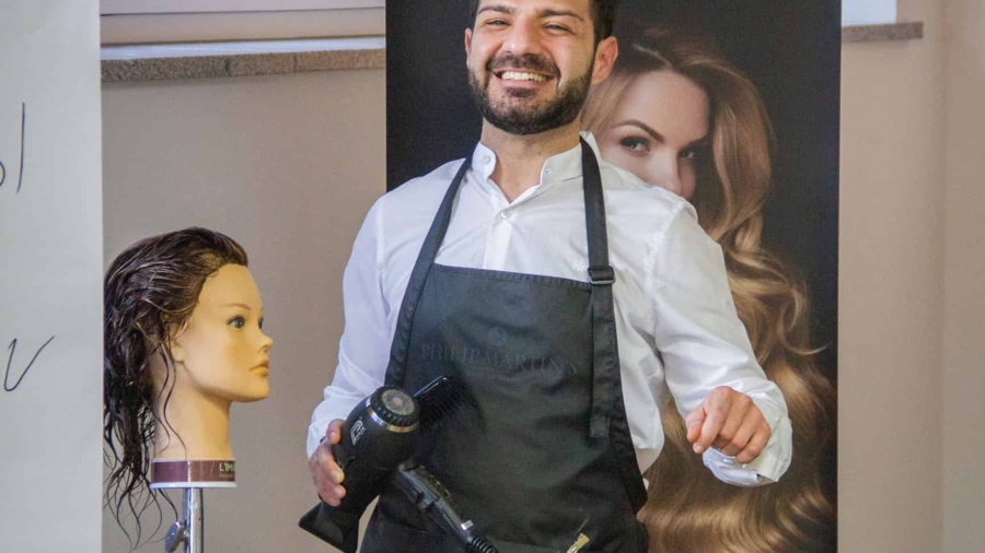 Friseur hält einen Haartrockner und steht neben einer Übungspuppe mit langen Haaren.