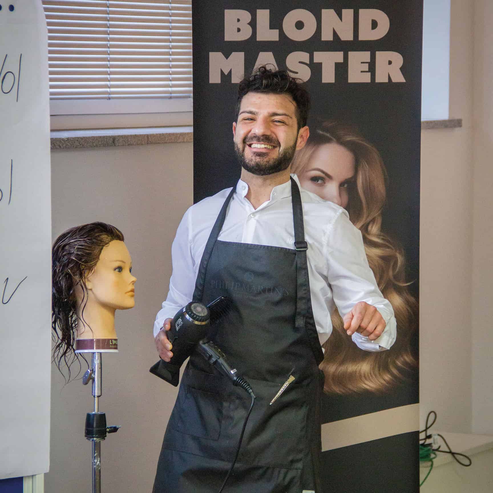 Friseur hält einen Haartrockner und steht neben einer Übungspuppe mit langen Haaren.