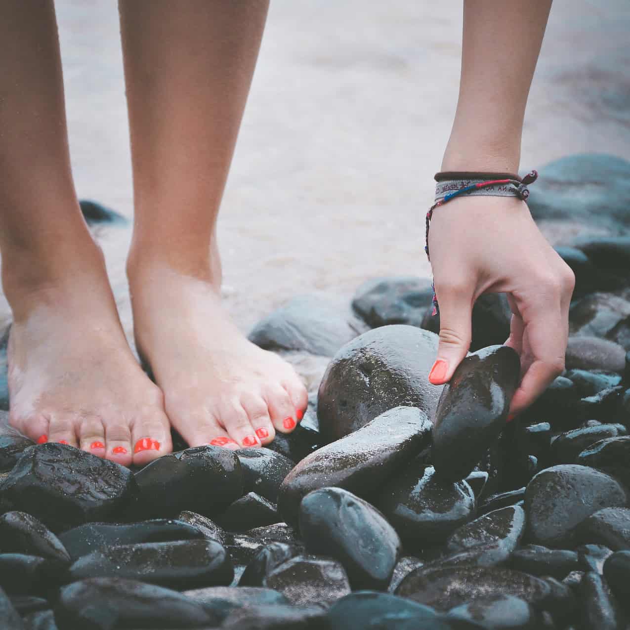 Eine Person mit nackten Füßen hebt einen glatten Stein von einem Kieselstrand auf.