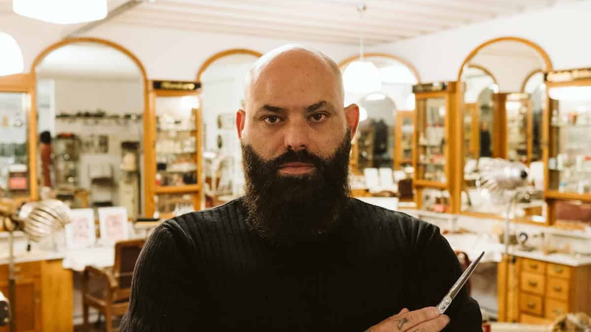 Ein Friseur mit Bart und Tätowierungen steht in einem Salon mit Holzregalen und Spiegeln.