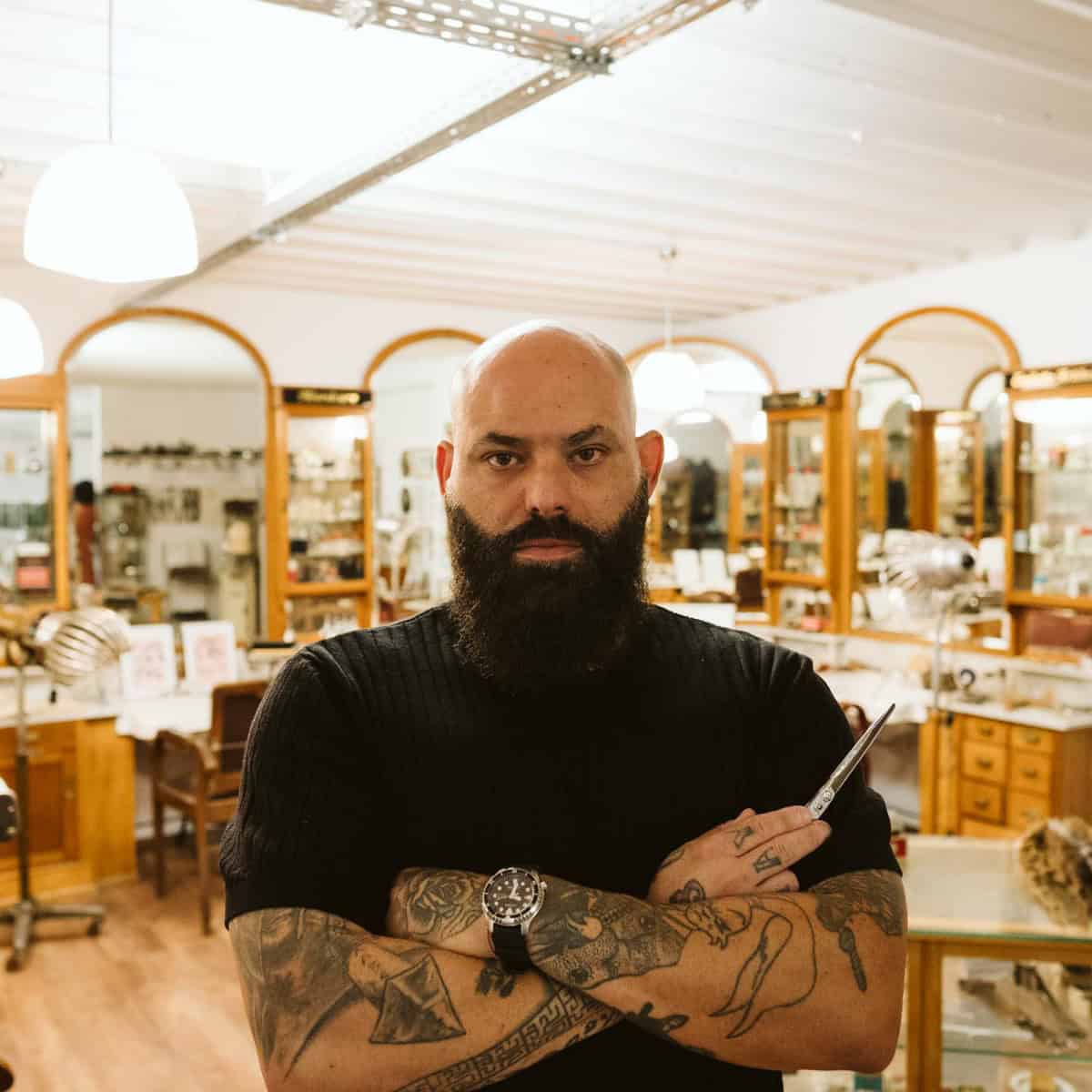 Ein Friseur mit Bart und Tätowierungen steht in einem Salon mit Holzregalen und Spiegeln.