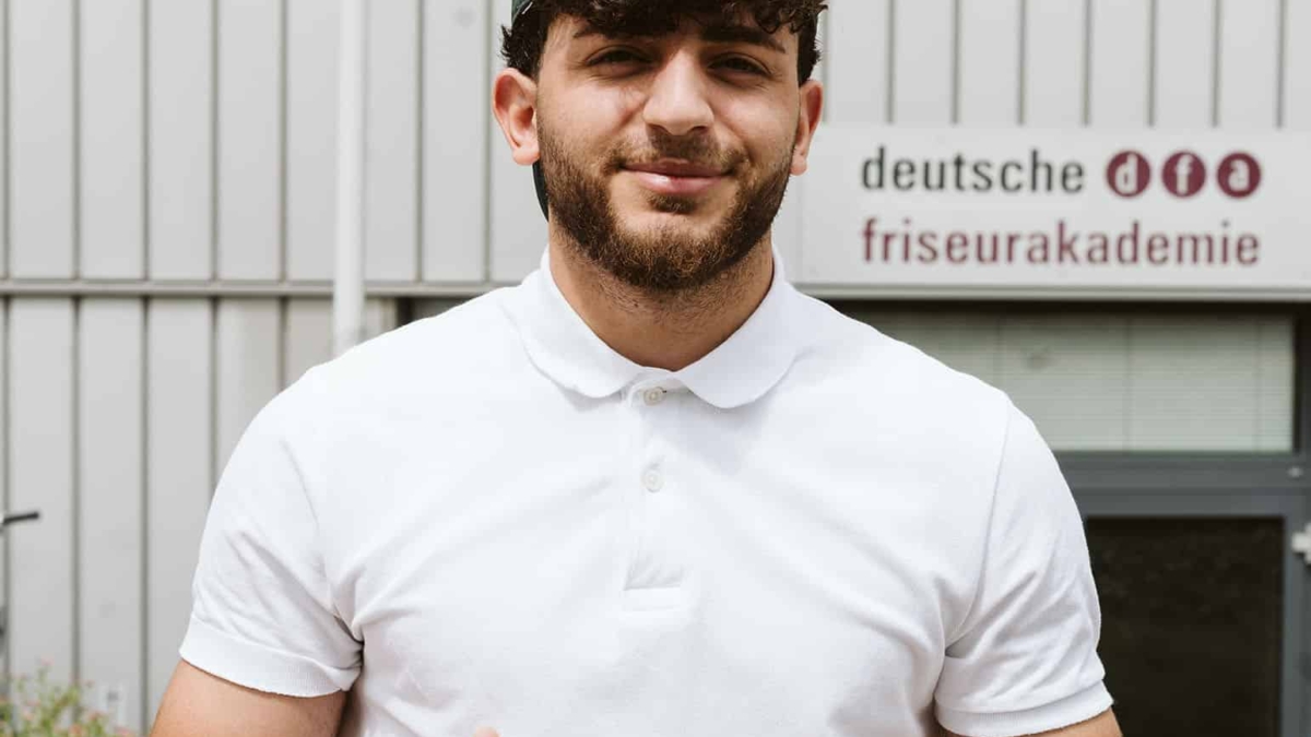 Junger Mann mit lockigem Haar und grüner Kappe zeigt Daumen hoch vor der Deutschen Friseurausbildung.