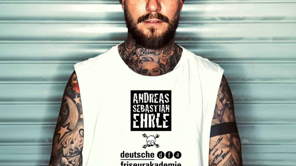 Mann mit Bart und Tattoos trägt ein weißes T-Shirt mit Aufdruck und Mütze.