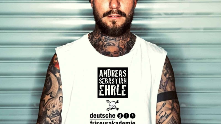 Mann mit Bart und Tattoos trägt ein weißes T-Shirt mit Aufdruck und Mütze.