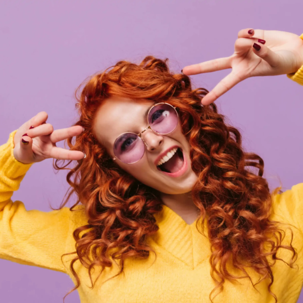 Eine Frau mit roter Dauerwelle und lila Sonnenbrille posiert vor lila Hintergrund in gelbem Pullover und zeigt lebendige Locken mit viel Volumen.