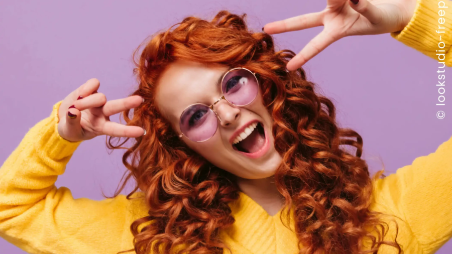 Eine Frau mit roter Dauerwelle und lila Sonnenbrille posiert vor lila Hintergrund in gelbem Pullover und zeigt lebendige Locken mit viel Volumen.