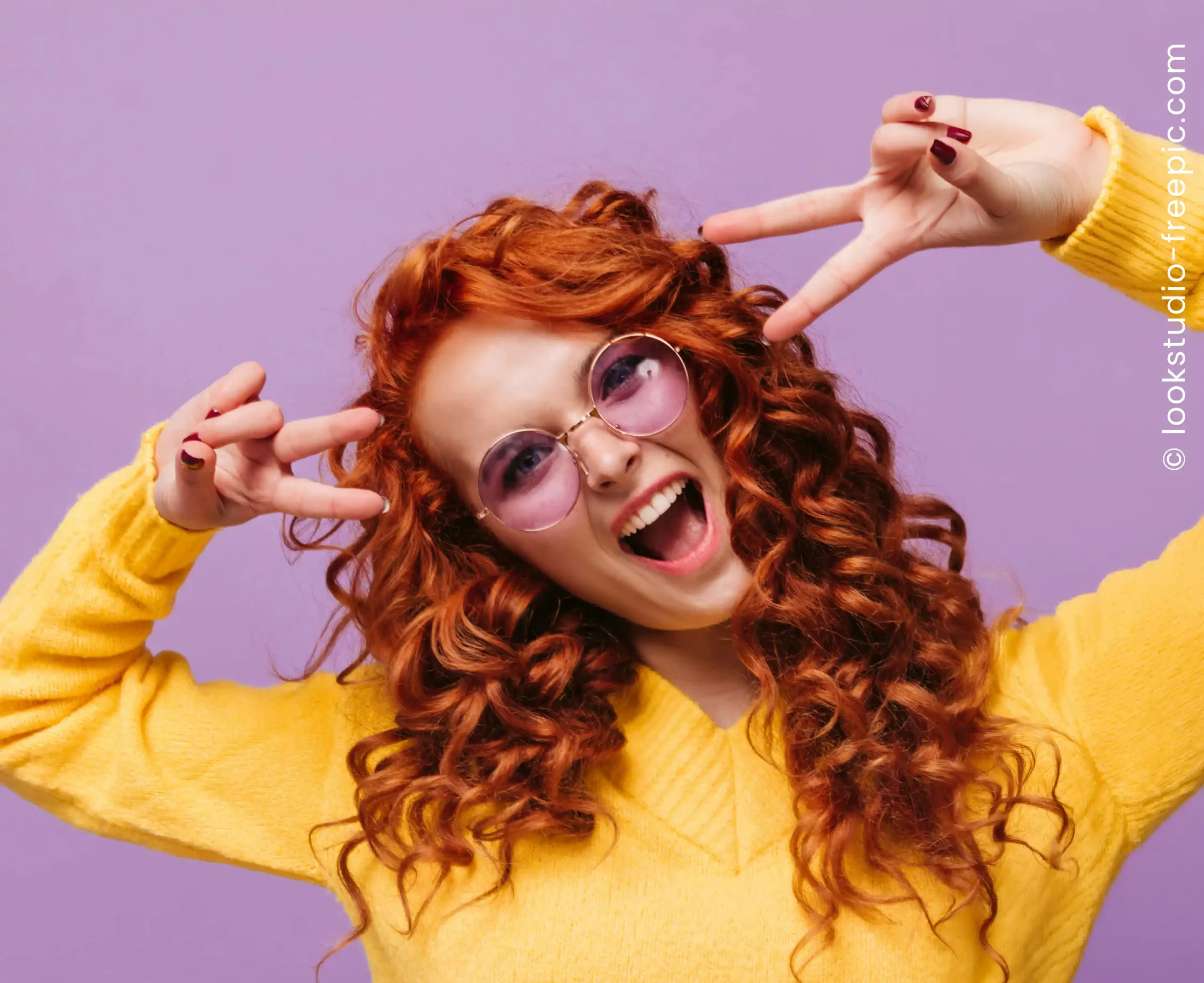 Eine Frau mit roter Dauerwelle und lila Sonnenbrille posiert vor lila Hintergrund in gelbem Pullover und zeigt lebendige Locken mit viel Volumen.